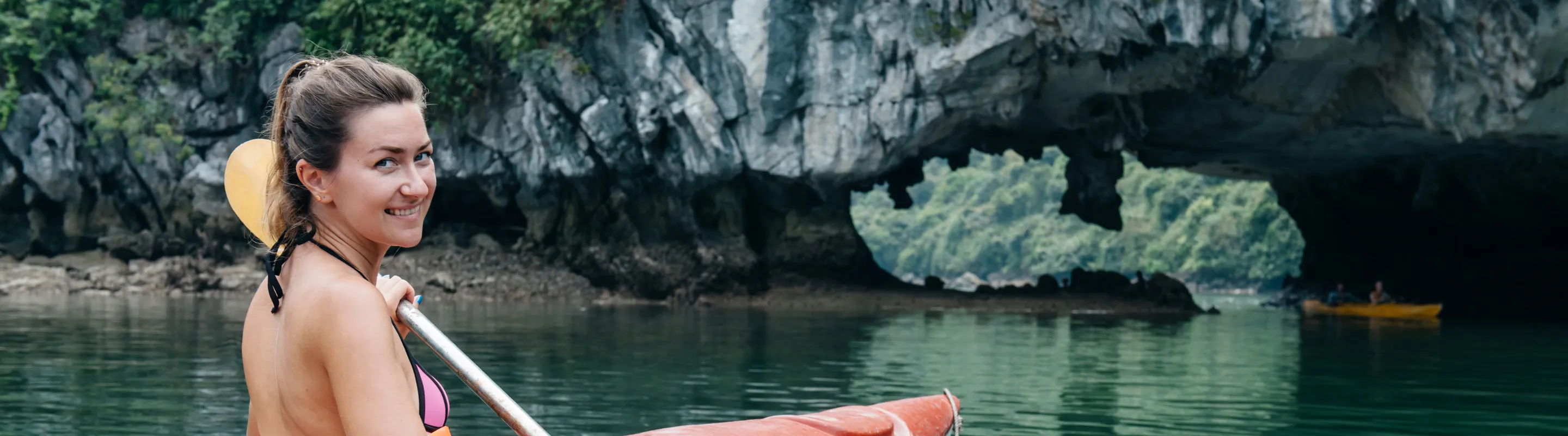 a woman with travel insurance for Vietnam paddles an orange kayak across calm green water near limestone cliffs