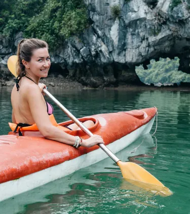a woman with travel insurance for Vietnam paddles an orange kayak across calm green water near limestone cliffs