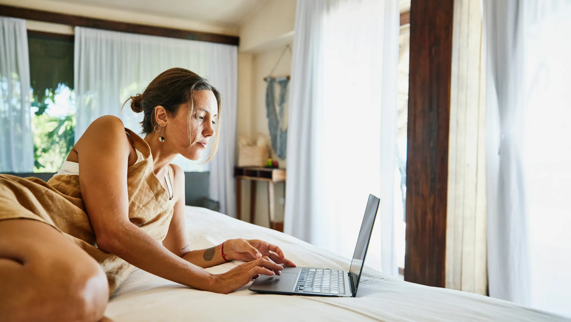 a woman looks at her laptop on the bed of her hotel room, researching travel insurance claim tips 