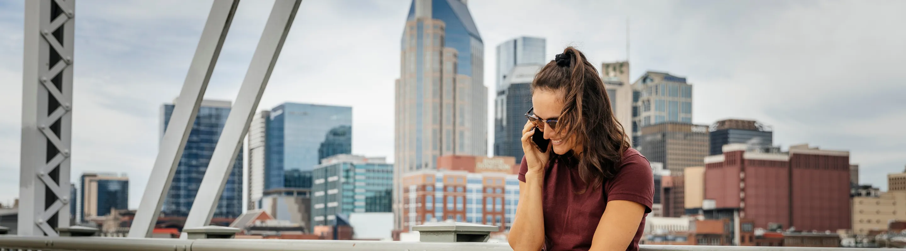 a woman stands talks on the phone on a bridge near the Nashville skyline, insured with travel insurance for Tennessee