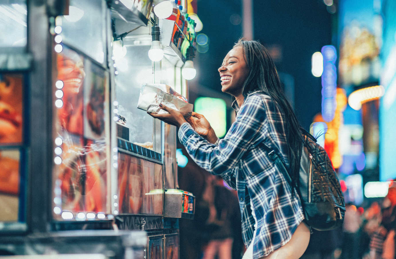a woman with travel insurance for New York smiles as she orders a hot dog in Times Square at night 