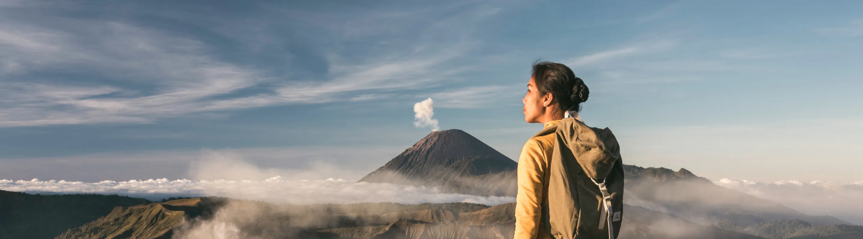 a woman with backpacker travel insurance stands on a grassy ridge overlooking a volcanic landscape with steam rising 