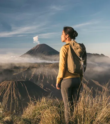 a woman with backpacker travel insurance stands on a grassy ridge overlooking a volcanic landscape with steam rising 