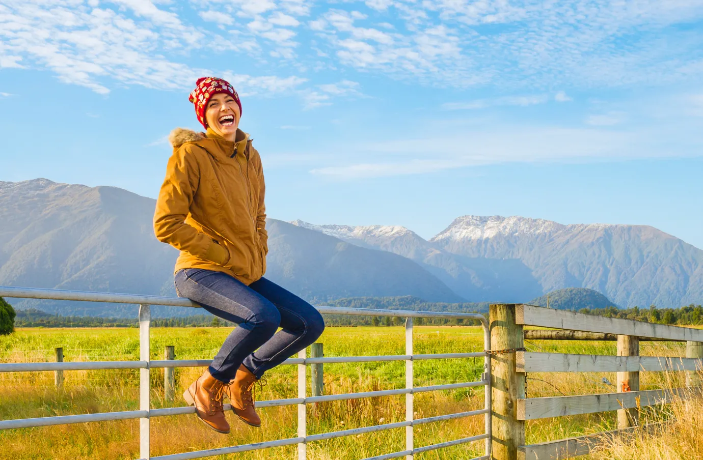 a woman in a yellow jacket sits on a fence near mountains, insured with travel insurance for New Zealand
