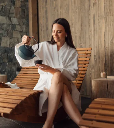 a woman in a spa robe sits on a wooden lounge chair, pouring tea, insured with travel insurance for wellness retreats