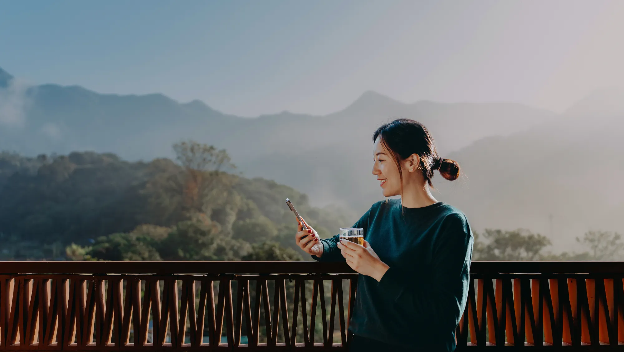 a woman looks at a travel app on her smartphone on a balcony with mountains in the background