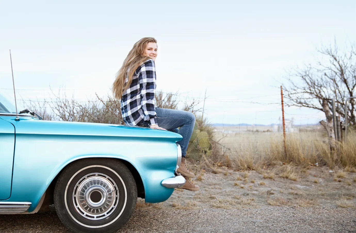 a young woman sits on a blue vintage car in the desert of West Texas, insured with travel insurance for Texas