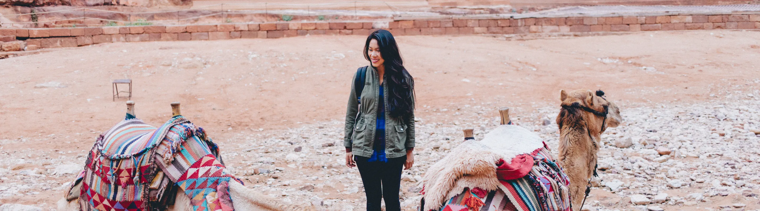 a young woman with travel insurance for Jordan cautiously stands by two camels in front of Petra