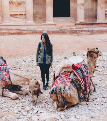 a young woman with travel insurance for Jordan cautiously stands by two camels in front of Petra