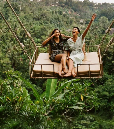 two women laugh on a giant jungle swing, insured with travel insurance for nature trips 