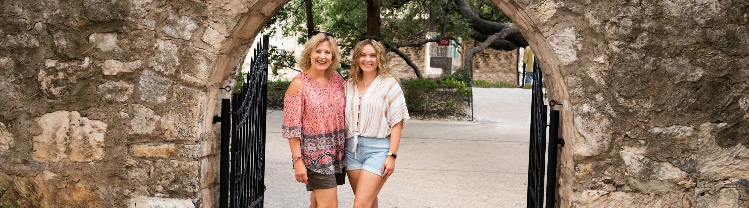 a mother-daughter duo with travel insurance for Texas stand under a stone archway by the Alamo in San Antonio
