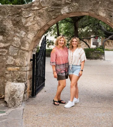 a mother-daughter duo with travel insurance for Texas stand under a stone archway by the Alamo in San Antonio