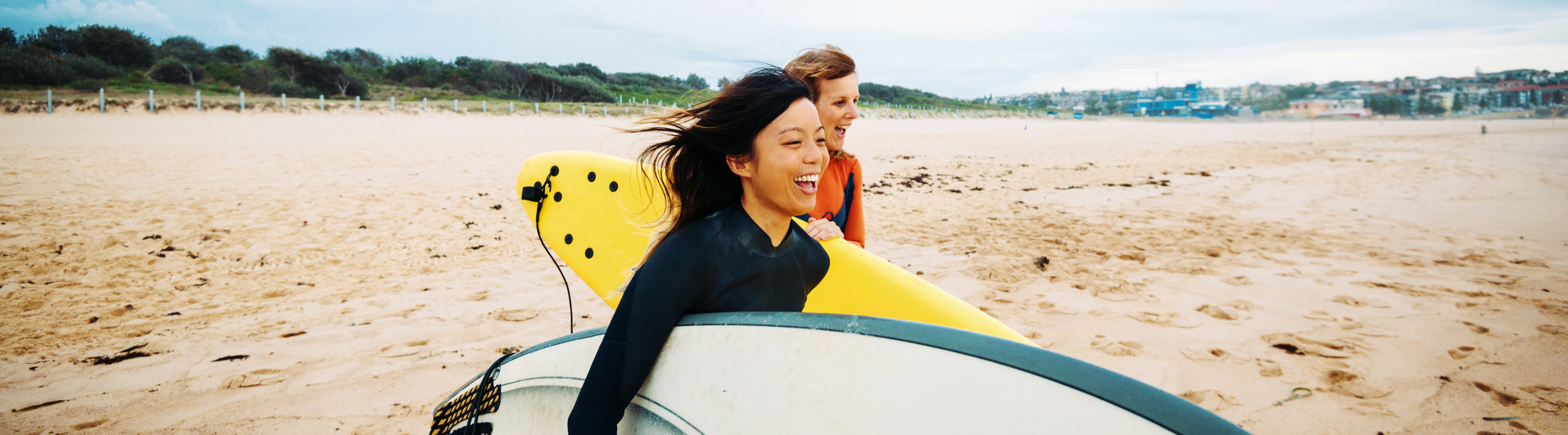 two women walk with their surfboards on a sandy shore, insured with travel insurance for Australia 