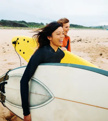 two women walk with their surfboards on a sandy shore, insured with travel insurance for Australia 