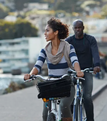 a couple with affordable travel insurance bicycles along a waterfront path near buildings and shoreline scenery