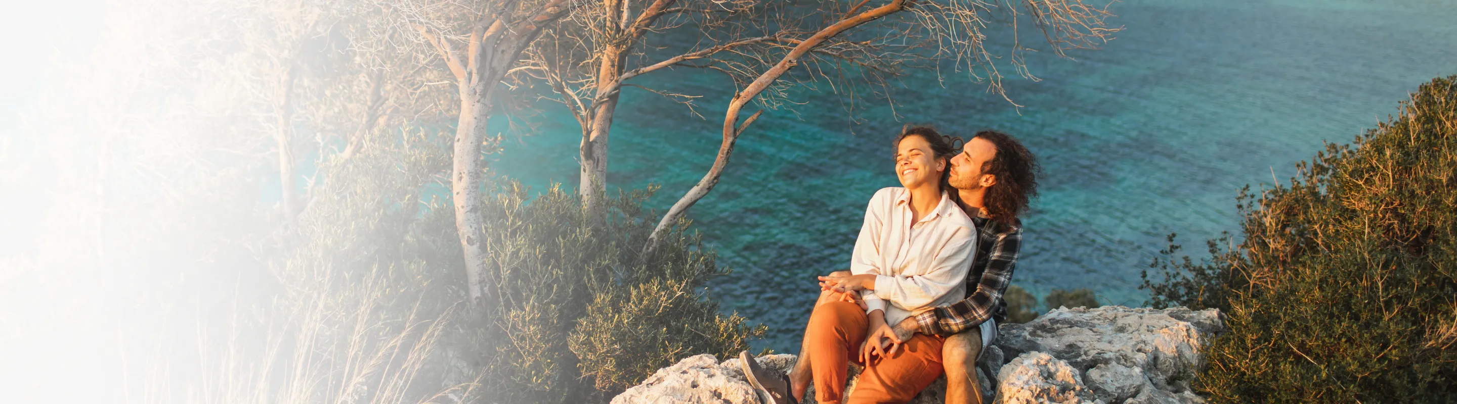 a young couple sits on rocky cliffs overlooking a turquoise bay surrounded by trees after reading about Travelex Insurance