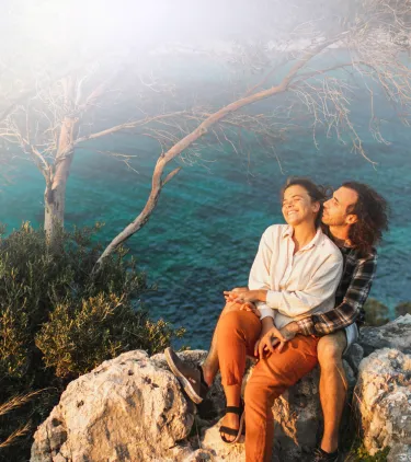 a young couple sits on rocky cliffs overlooking a turquoise bay surrounded by trees after reading about Travelex Insurance