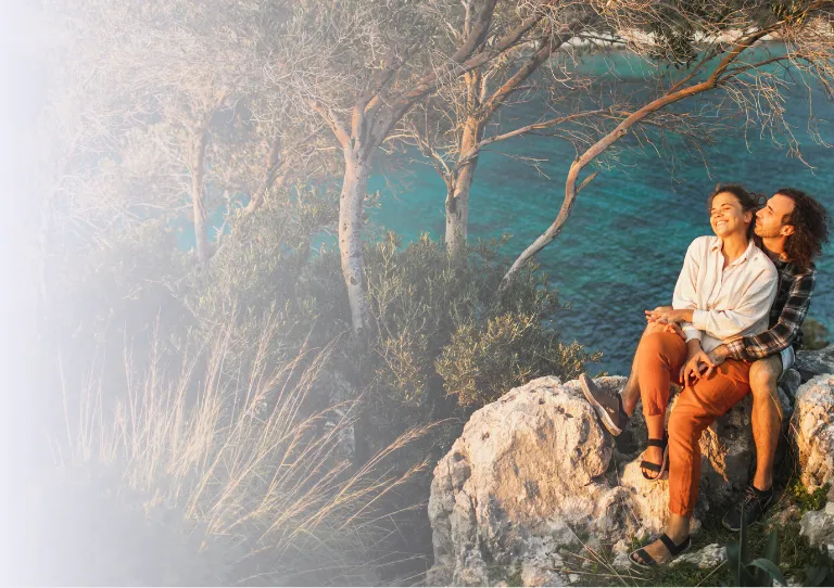 a young couple sits on rocky cliffs overlooking a turquoise bay surrounded by trees after reading about Travelex Insurance