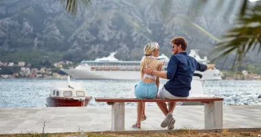 A smiling couple on a bench with a cruise ship in the background, which may have them wondering, &quot;How safe is cruising?&quot;.