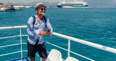 Smiling man searching how to get the best deal on a cruise on his phone with a ship in background.
