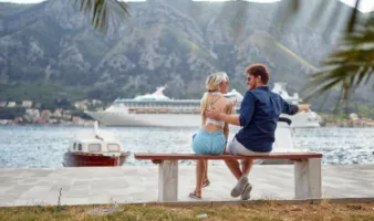 A smiling couple on a bench with a cruise ship in the background, which may have them wondering, &quot;How safe is cruising?&quot;.