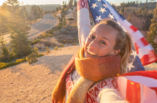 Young woman smiling and taking a selfie while holding an American flag, highlighting the use of a United States travel guide